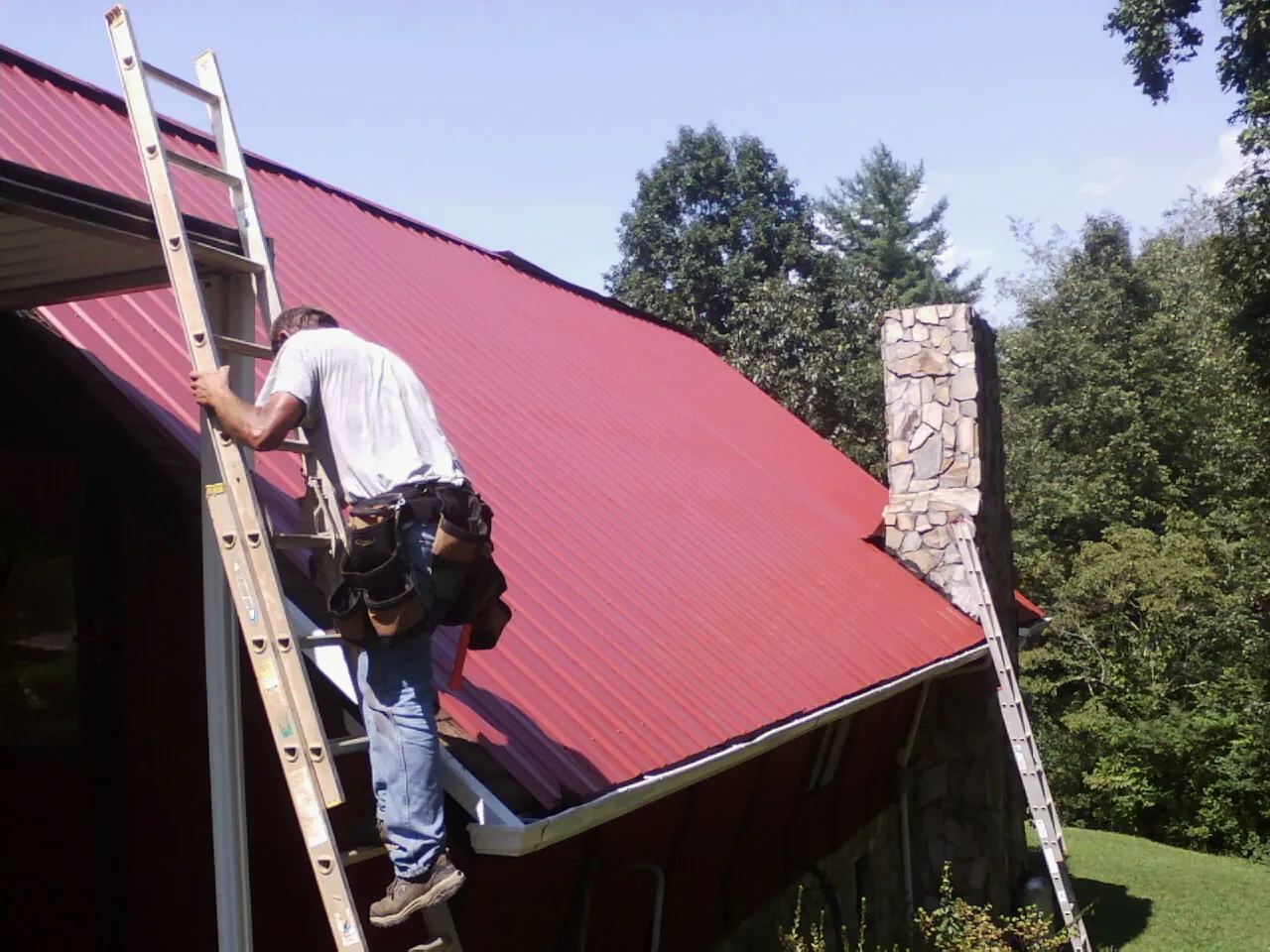 Roofer on a ladder beside a red metal roof and stone chimney — Soffit & Fascia Repair by Roofing Brothers in Waupun